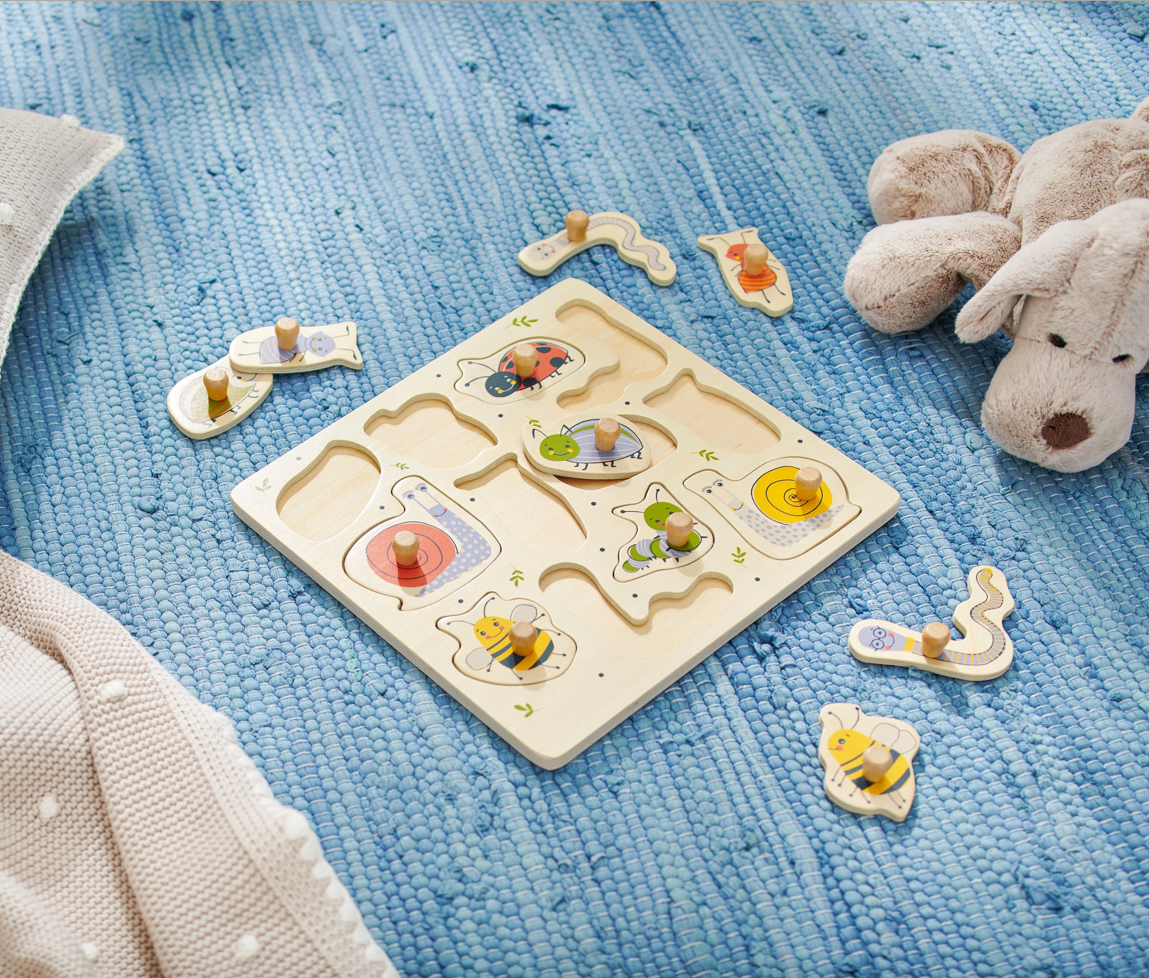 Dřevěné puzzle pro nejmenší s motivy hmyzu leží na modrém koberci vedle plyšového polštáře a hračky.