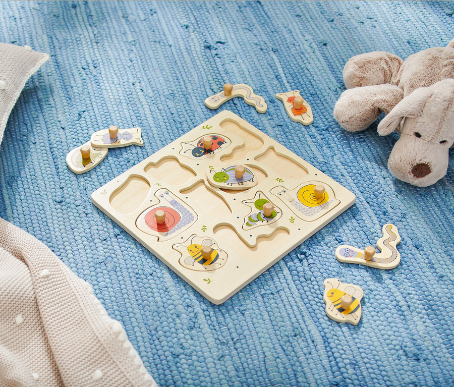 Dřevěné puzzle pro nejmenší s motivy hmyzu leží na modrém koberci vedle plyšového polštáře a hračky.