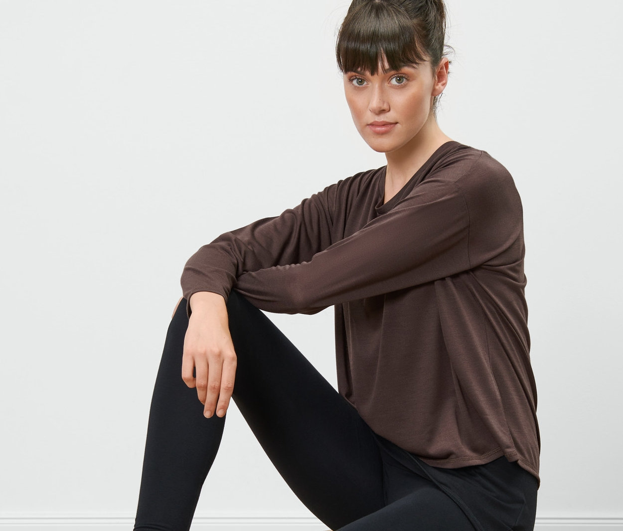 Žena sedí na dřevěné stoličce a má na sobě hnědé sportovní triko 2 v 1 a černé legíny.