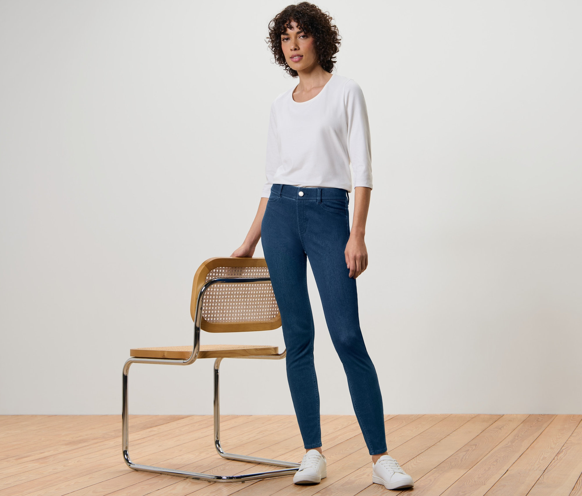 Žena pózuje vedle židle a má na sobě džegíny, bílou blůzu a tenisky.