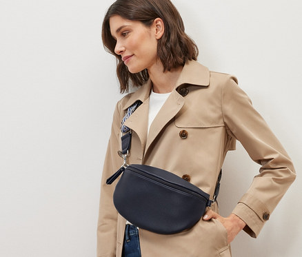 Žena v béžovém trenčkotu a džínech pózuje s crossbody taškou.