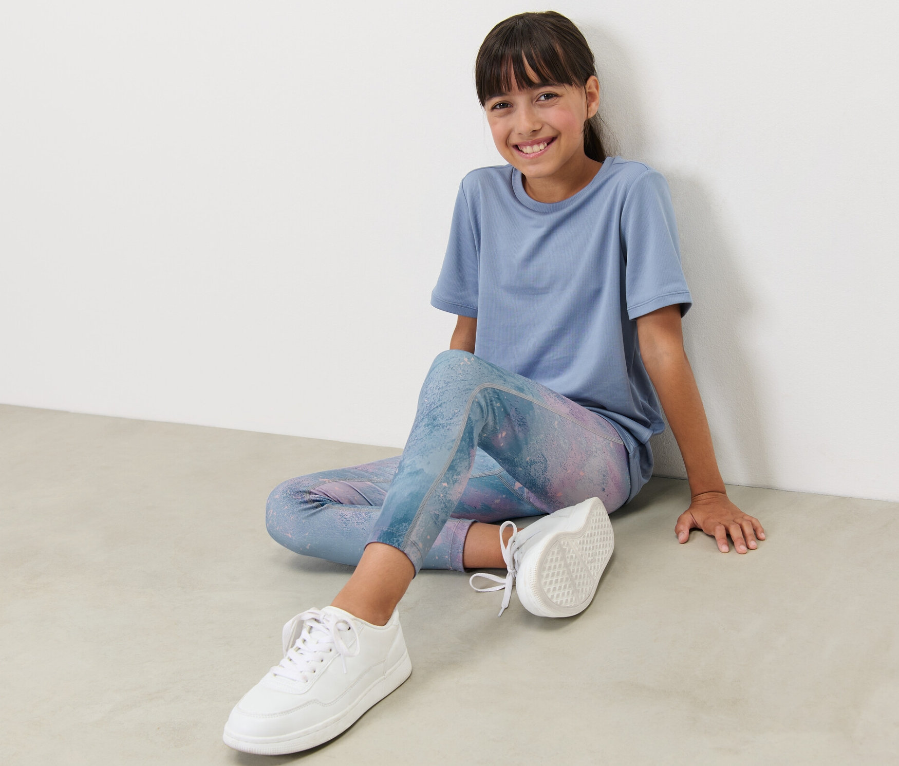 Dívka sedí na zemi a má na sobě dětské sportovní legíny s potiskem a modré tričko.