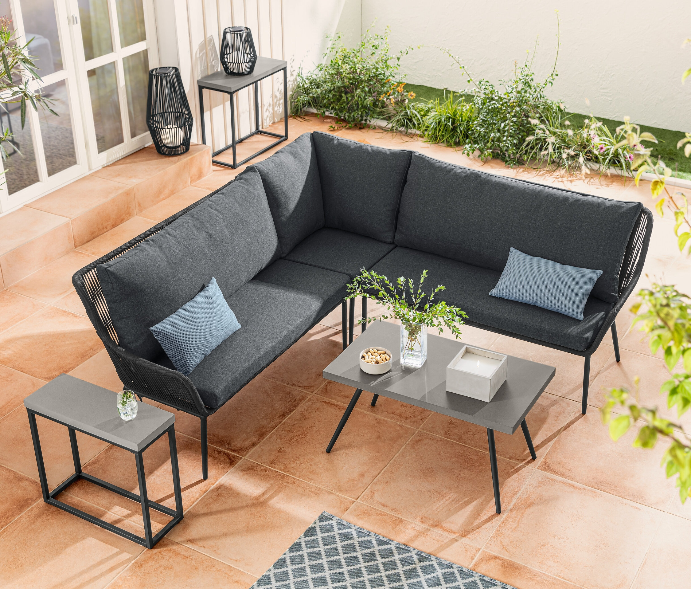 Rohová sedací souprava s textilním výpletem stojí vedle konferenčního stolku s polycementovou deskou. Sada konzolových stolků s polycementovou deskou je umístěna vedle solární lucerny s textilním výpletem.