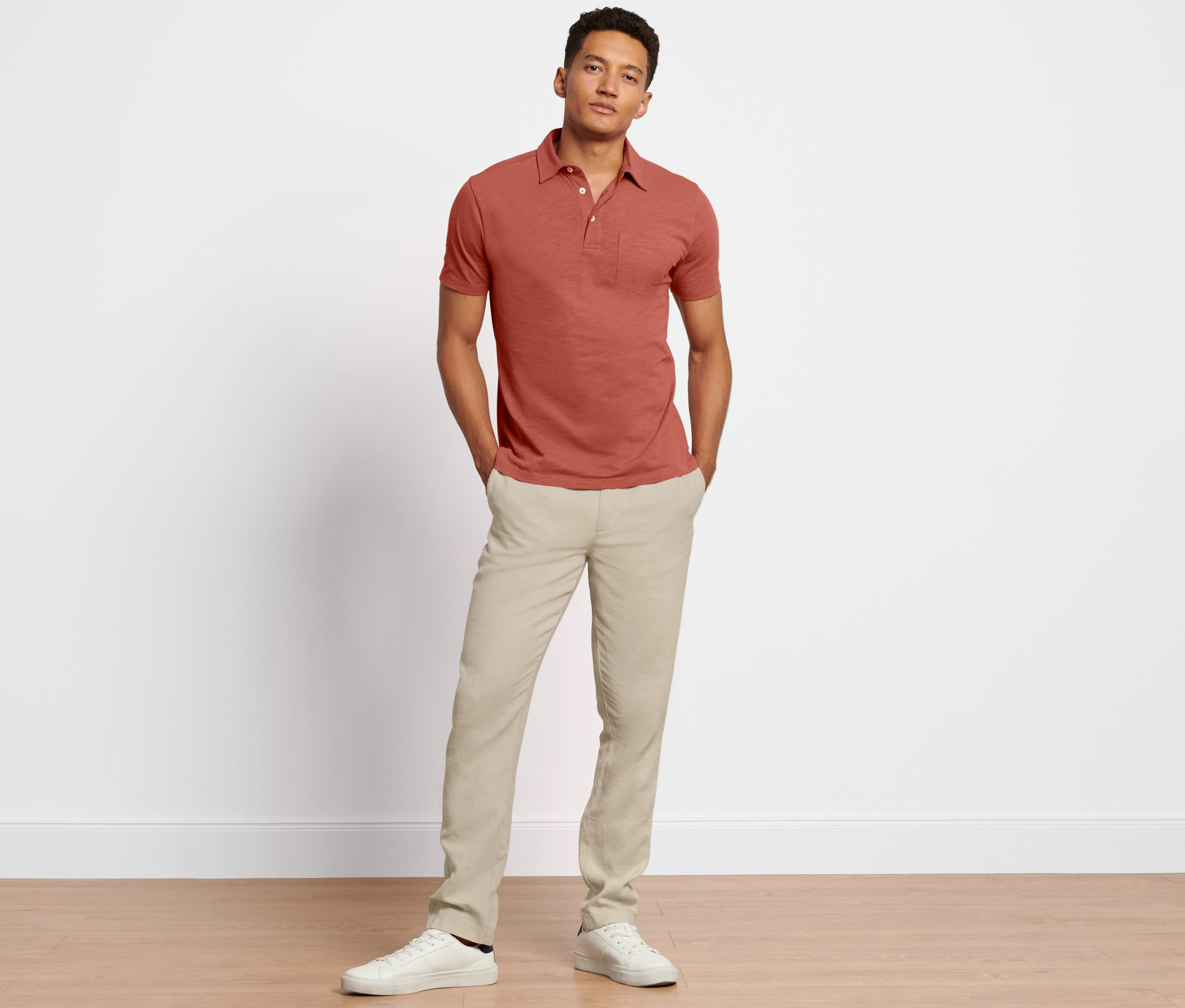 Muž stojí a pózuje v lněných kalhotách chino a červenohnědé košili polo.