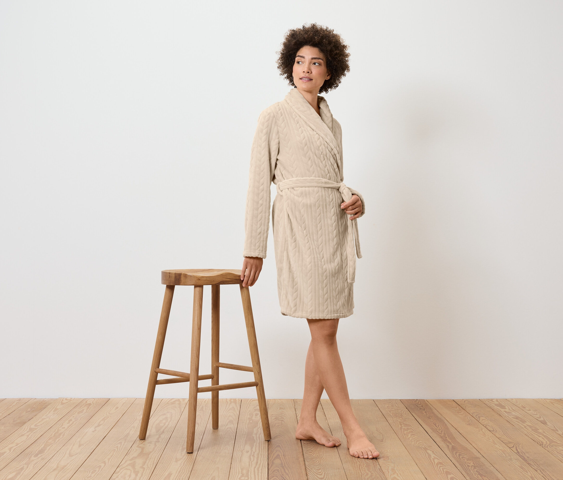 Žena v krémovém županu stojí vedle dřevěné stoličky.