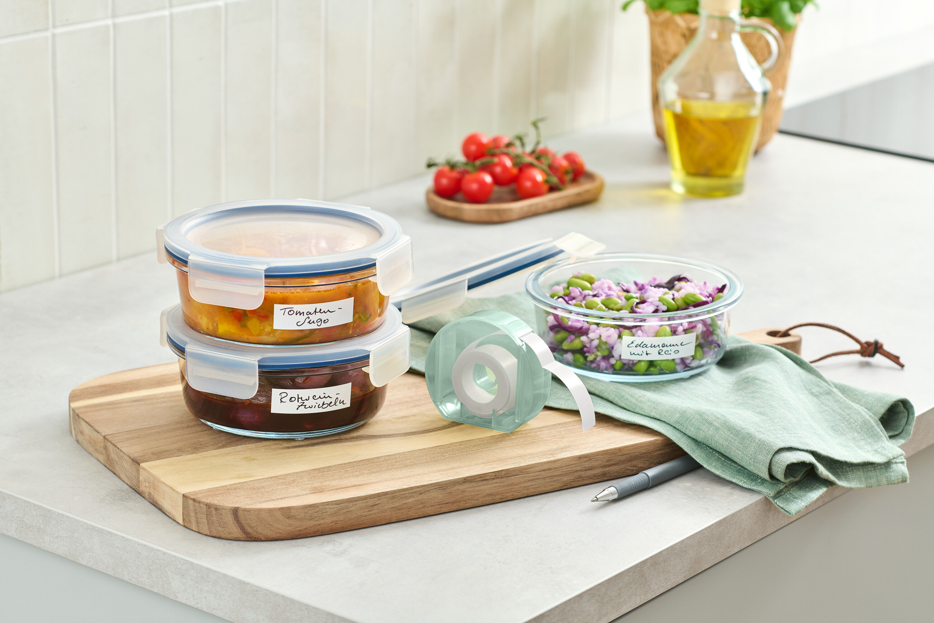 Dva skleněné zásobníky s jídlem naskládané na dřevěném prkénku vedle role s etiketami.