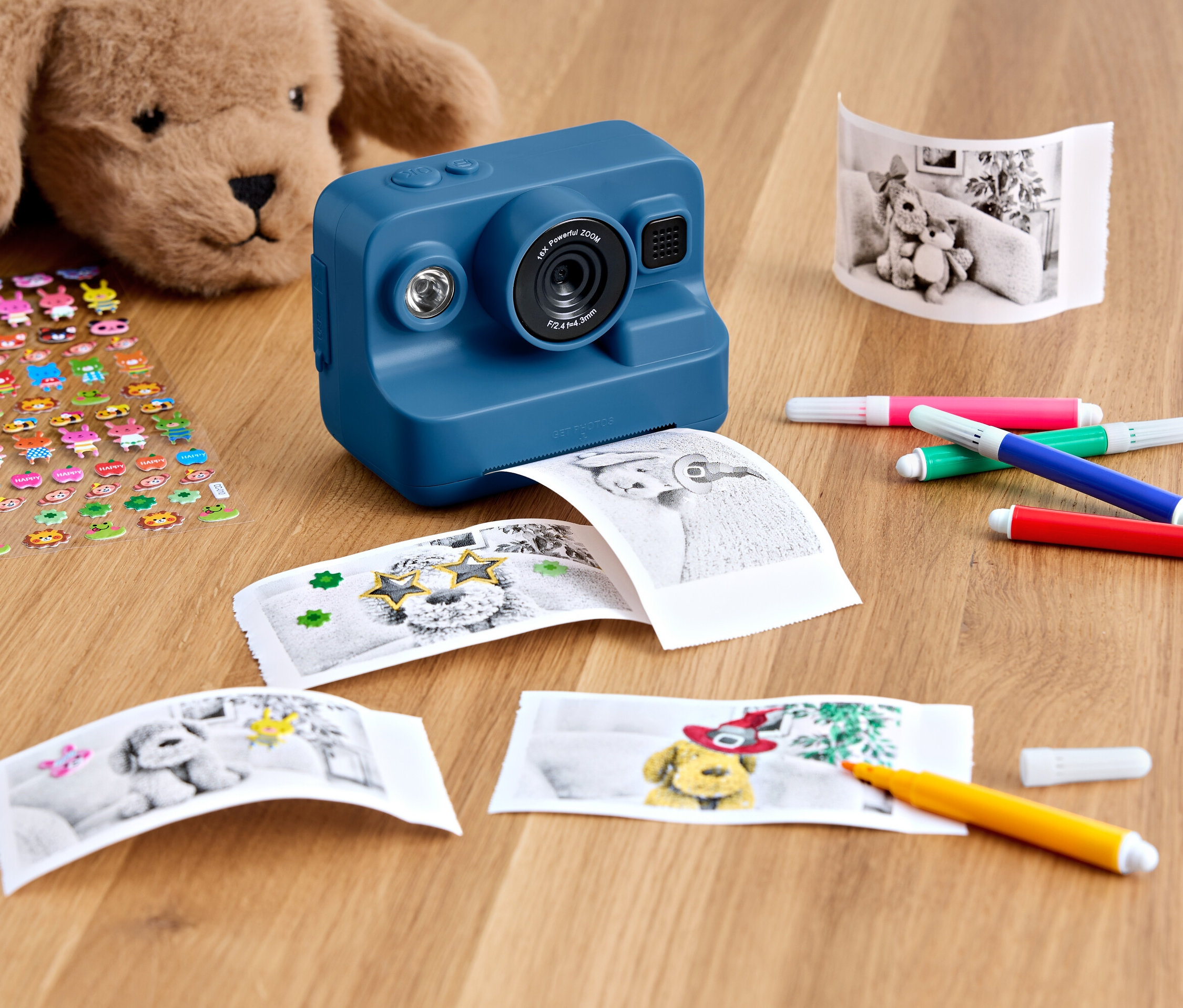 Modrá tiskací kamera leží na dřevěném stole s plyšovým medvídkem, archem samolepek, vytištěnými fotografiemi a barevnými fixami.