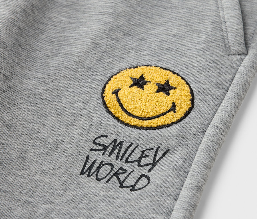 Detail šedých dětských joggingových kalhot SMILEYWORLD® s nášivkou žlutého smajlíka.