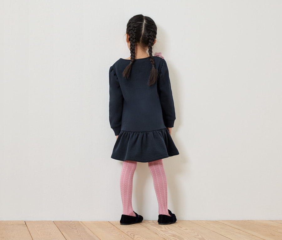 Dívka s copánky stojí zády, má na sobě modré šaty, růžové vzorované punčocháče a černé boty.