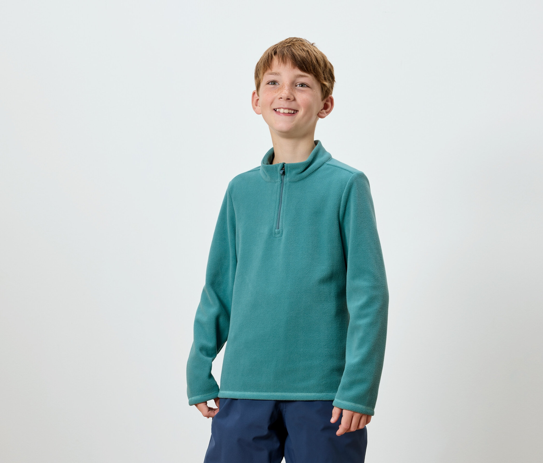 Usmívající se chlapec pózuje v tyrkysové mikině na zip a tmavě modrých kalhotách.