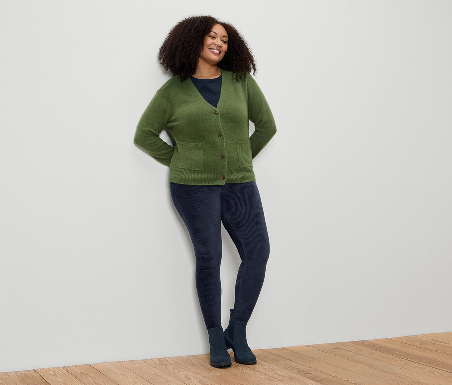 Žena s kudrnatými vlasy se opírá o zeď. Má na sobě zelený kardigan, modré legíny a boty.