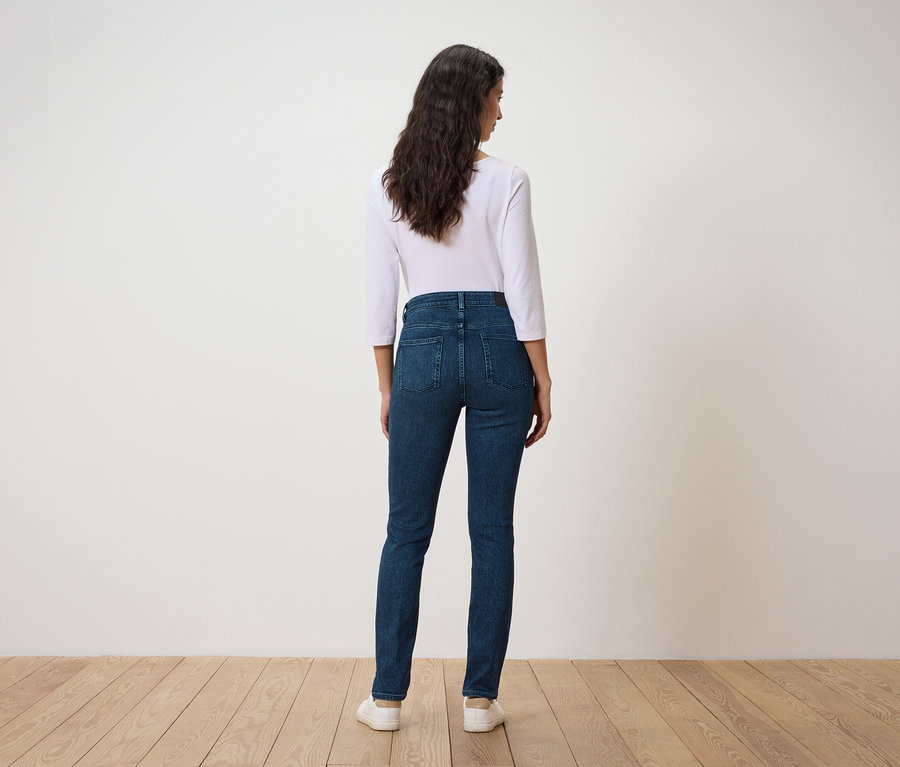 Žena s tmavými vlnitými vlasy stojí zády, oblečená do bílého trička s tříčtvrtečním rukávem a tmavě modrých džínů.