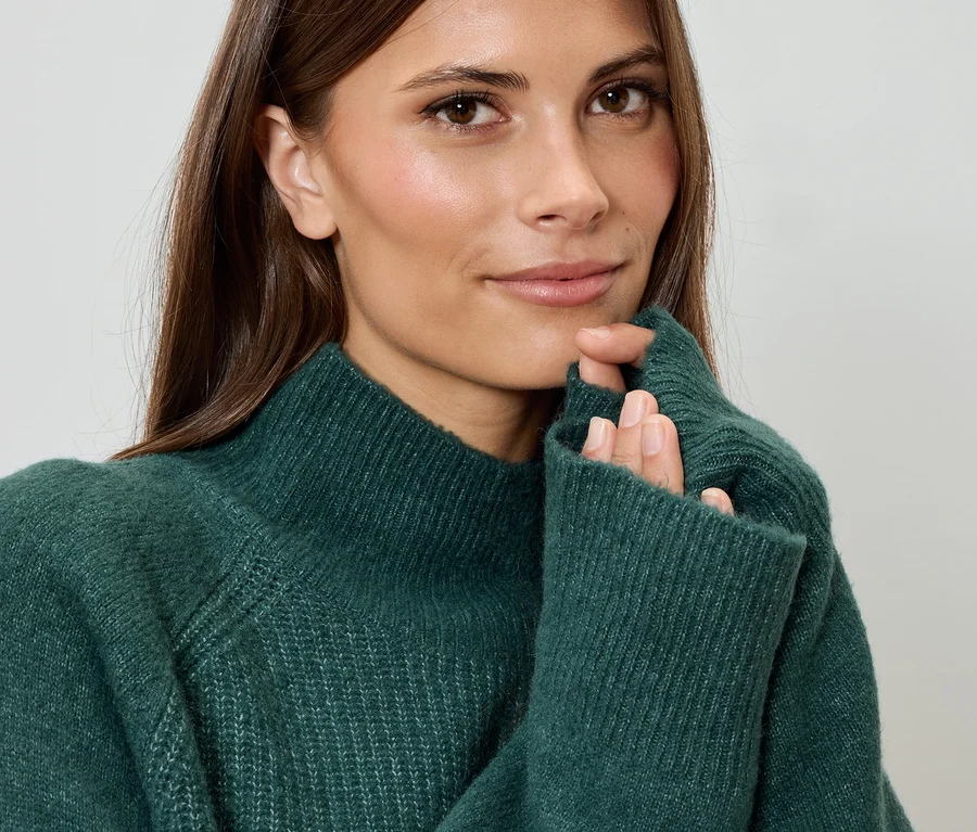 Žena se dívá do kamery a má ruce u obličeje. Oblečená je do zeleného svetru.