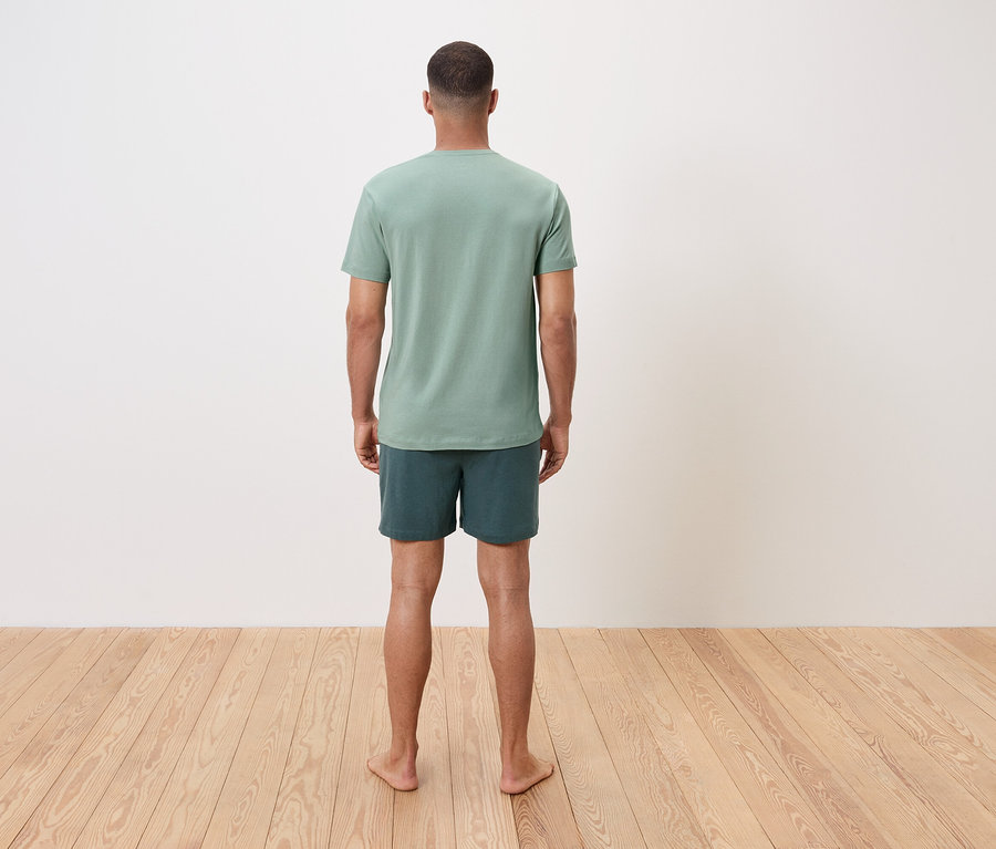 Muž stojí bosý zády k divákovi, má na sobě zelené tričko a šortky.