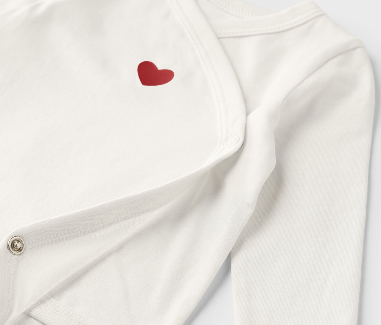 Detail bílého rostoucího body pro miminka s červeným srdíčkem.