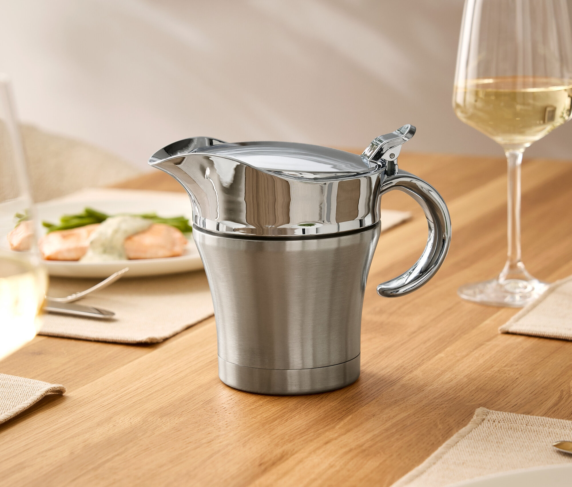 Na dřevěném stole stojí nerezová omáčník s pokličkou, sklenice vína a talíře s jídlem.