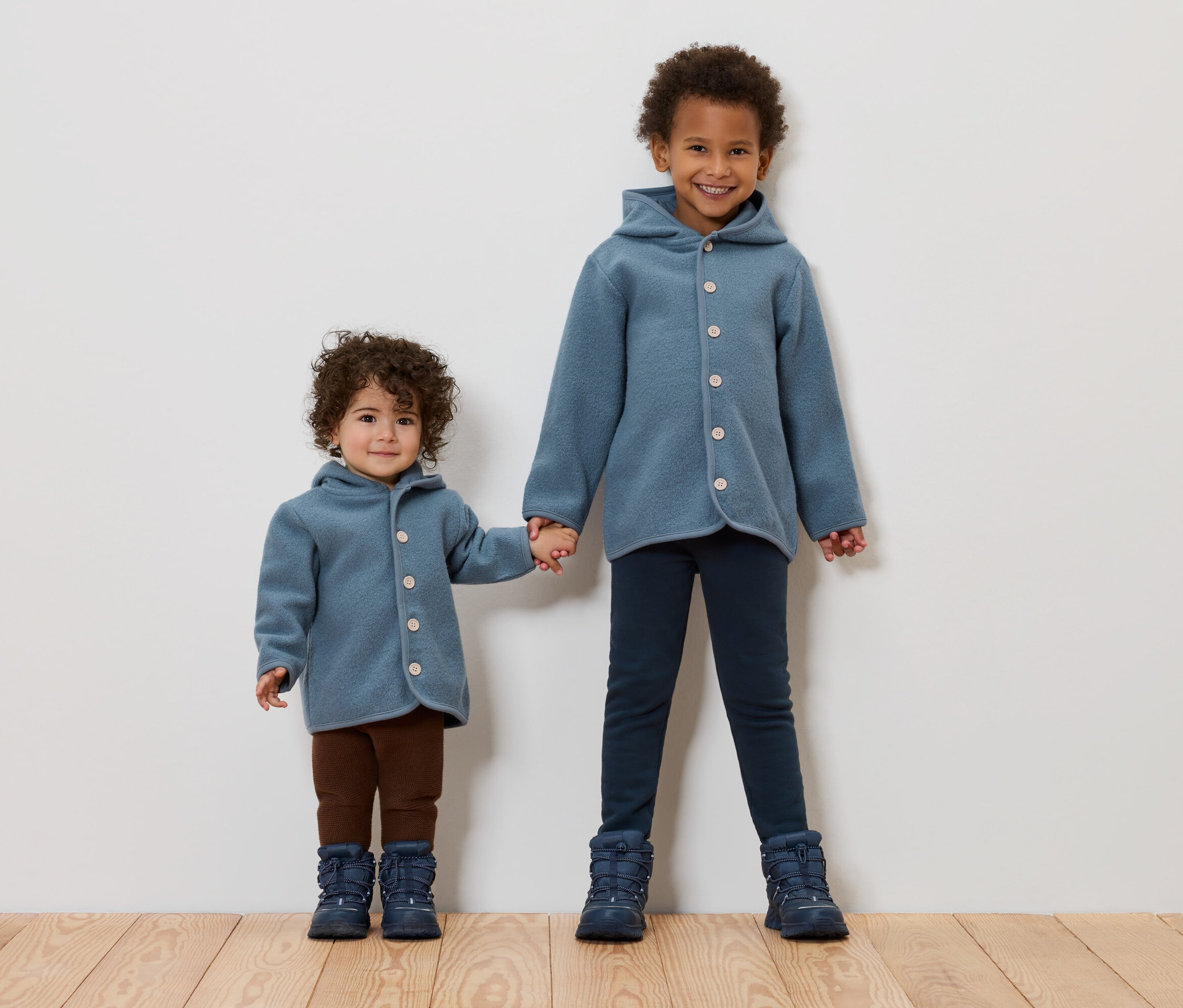 Dvě děti drží se za ruce. Obě mají na sobě modré kabáty a boty.