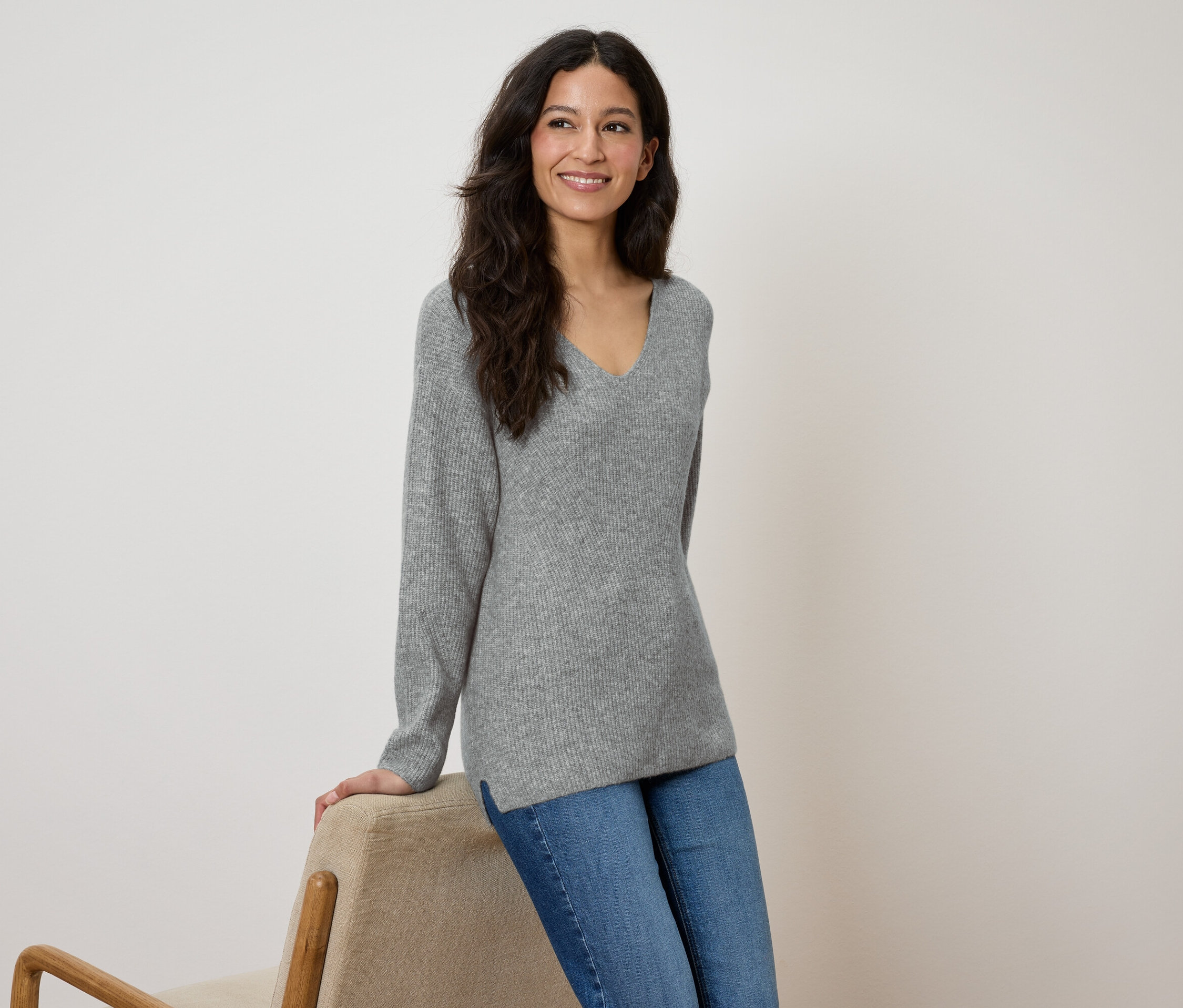 Žena se usmívá a opírá se o béžovou židli, má na sobě šedý svetr a modré džíny.