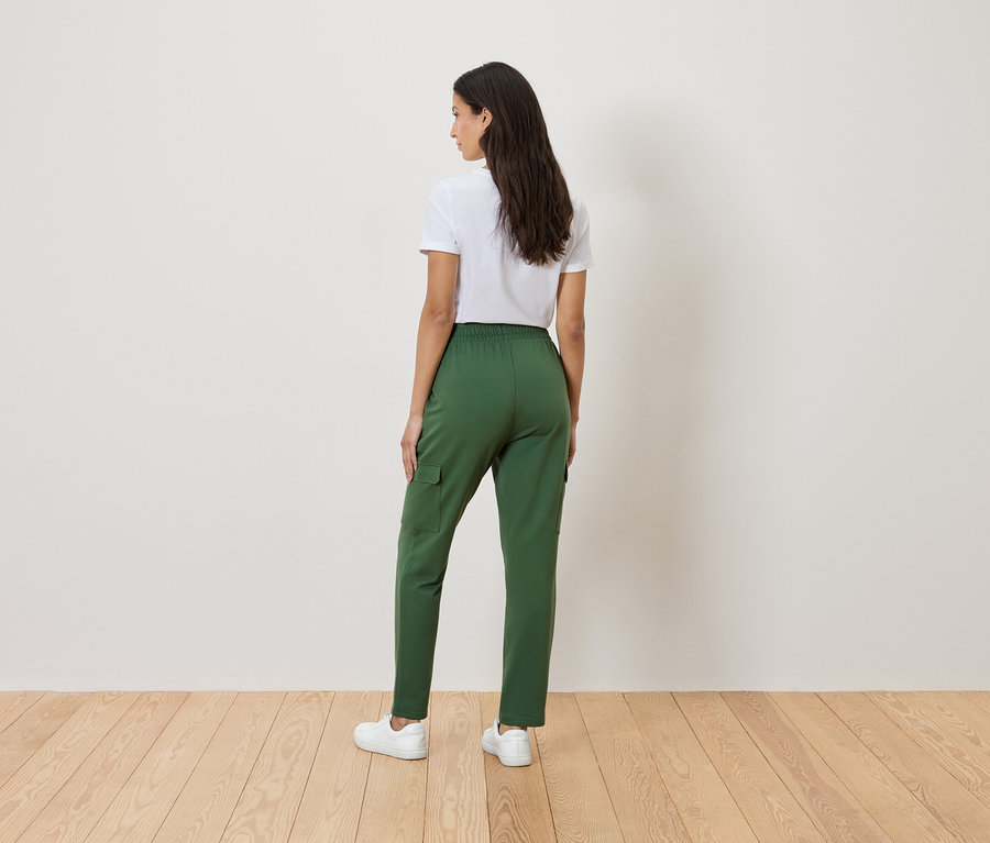 Žena stojí zády ke kameře a má na sobě bílé tričko, zelené kalhoty a bílé tenisky.