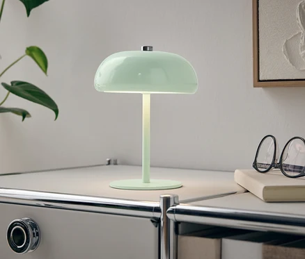 Zelená lampa stojí na bílém nočním stolku vedle rostliny a brýlí.