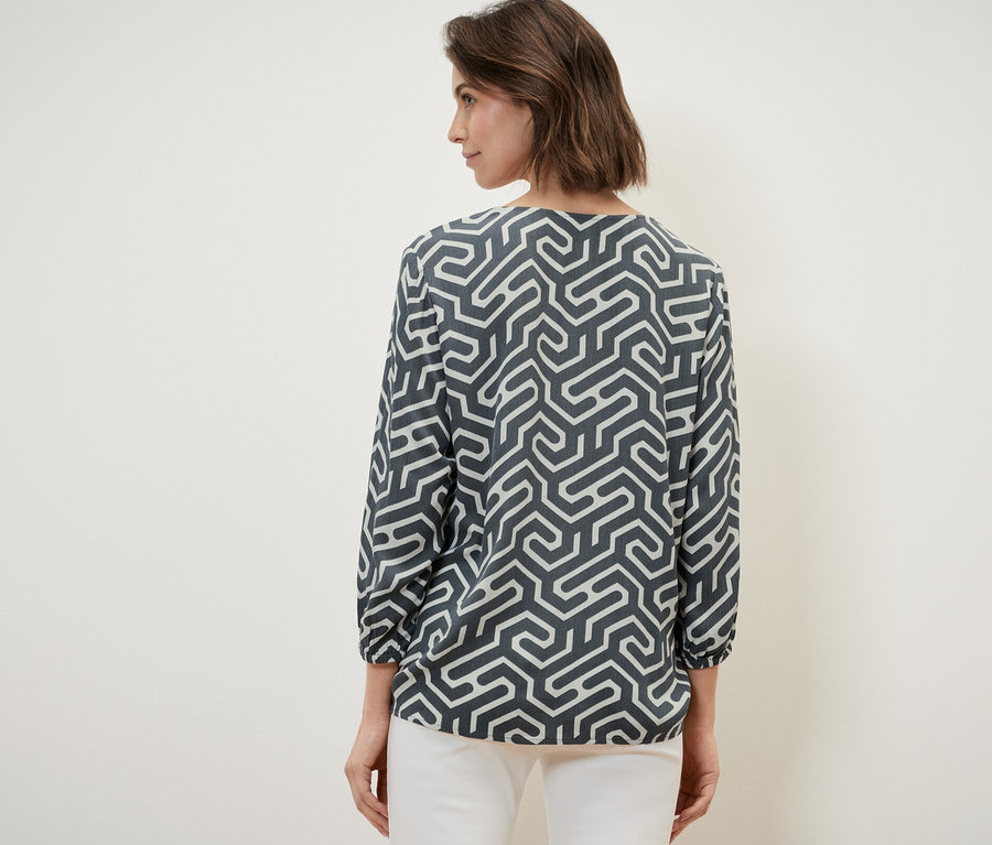 Žena zády k fotoaparátu má na sobě halenku v mačkaném vzhledu a bílé kalhoty.