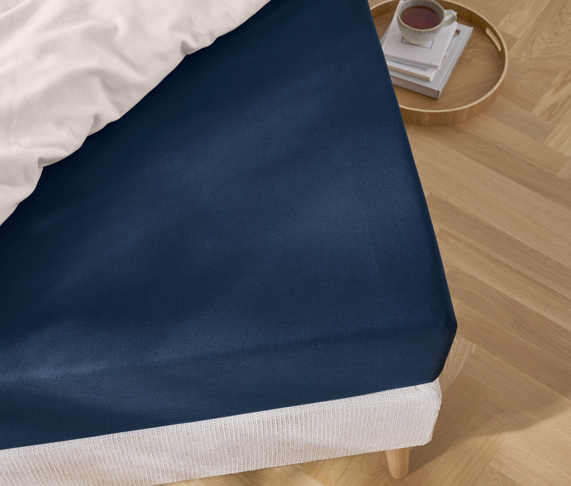 Detail modrého prostěradla na posteli s bílými peřinami a tácem s knihami a šálkem.