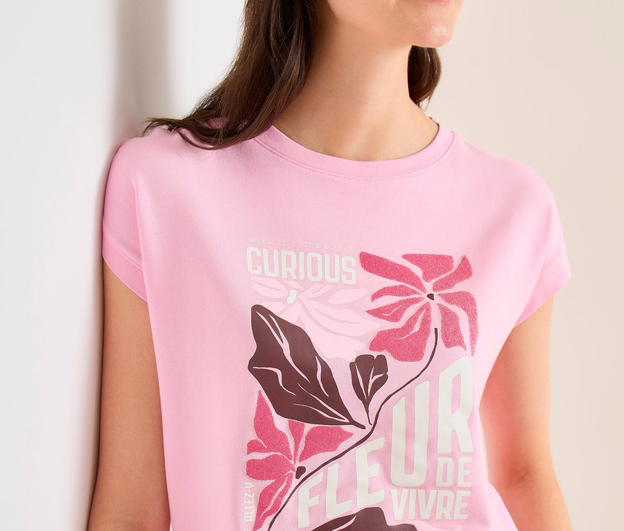Žena v béžové kšiltovce a růžovém Tričku Placement CECIL s květinovým vzorem a nápisem "FLEUR DE VIVRE" se opírá o zeď.