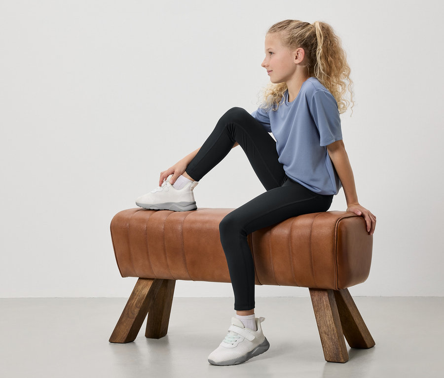 Mladá dívka s blond vlasy svázanými do culíku sedí na gymnastickém koni. Má na sobě modré tričko, černé legíny a bílé tenisky.