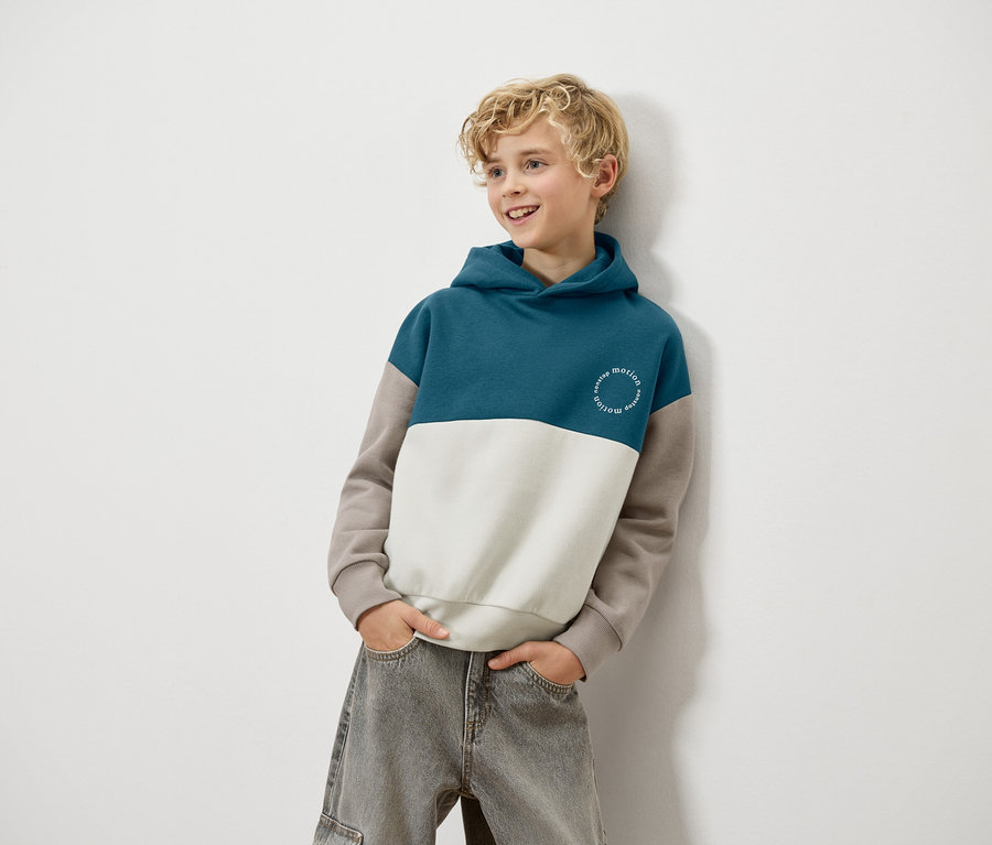 Blonďatý chlapec se opírá o zeď a má na sobě modro-bílo-hnědou mikinu s kapucí a šedé džíny.