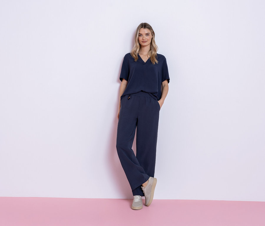 Žena stojí na růžovém a bílém pozadí a má na sobě tmavě modrý outfit skládající se z trička a kalhot a šedé tenisky.