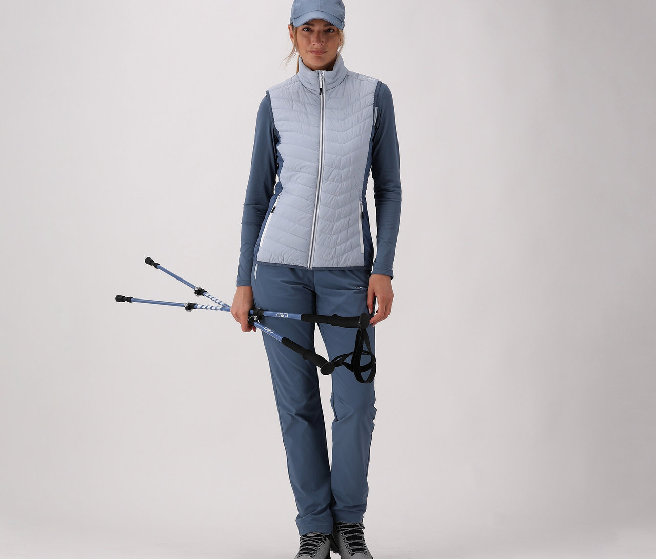 Žena v modrém sportovním oblečení s dámskou hybridní vestou CMP drží trekové hole na světlém pozadí.