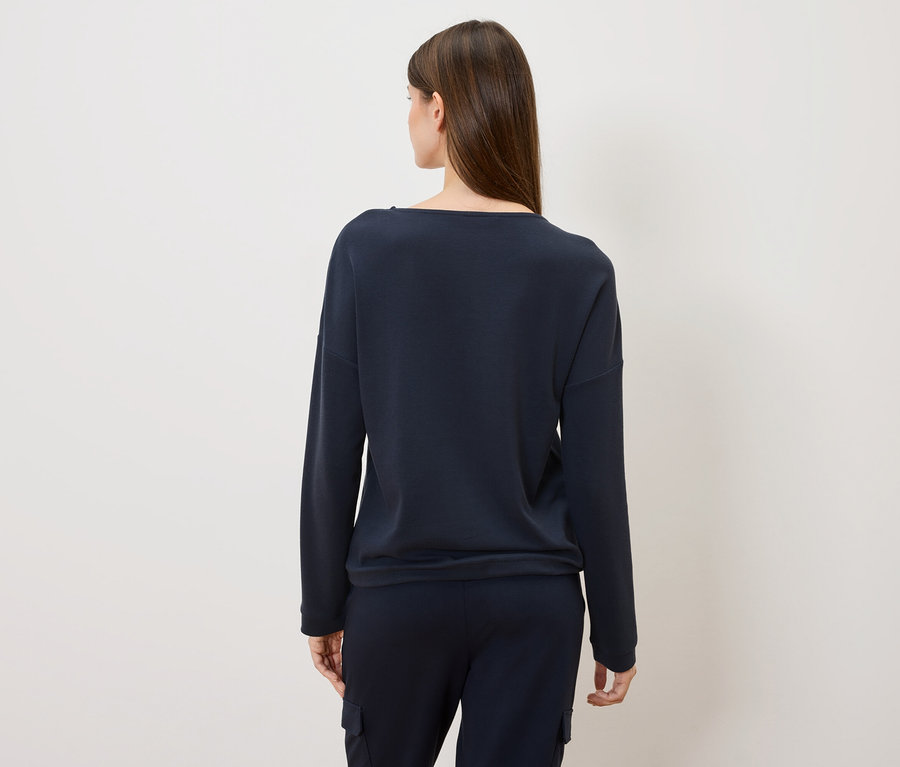 Pohled zezadu na ženu s dlouhými hnědými vlasy v tmavě modrém svetru a kalhotách.