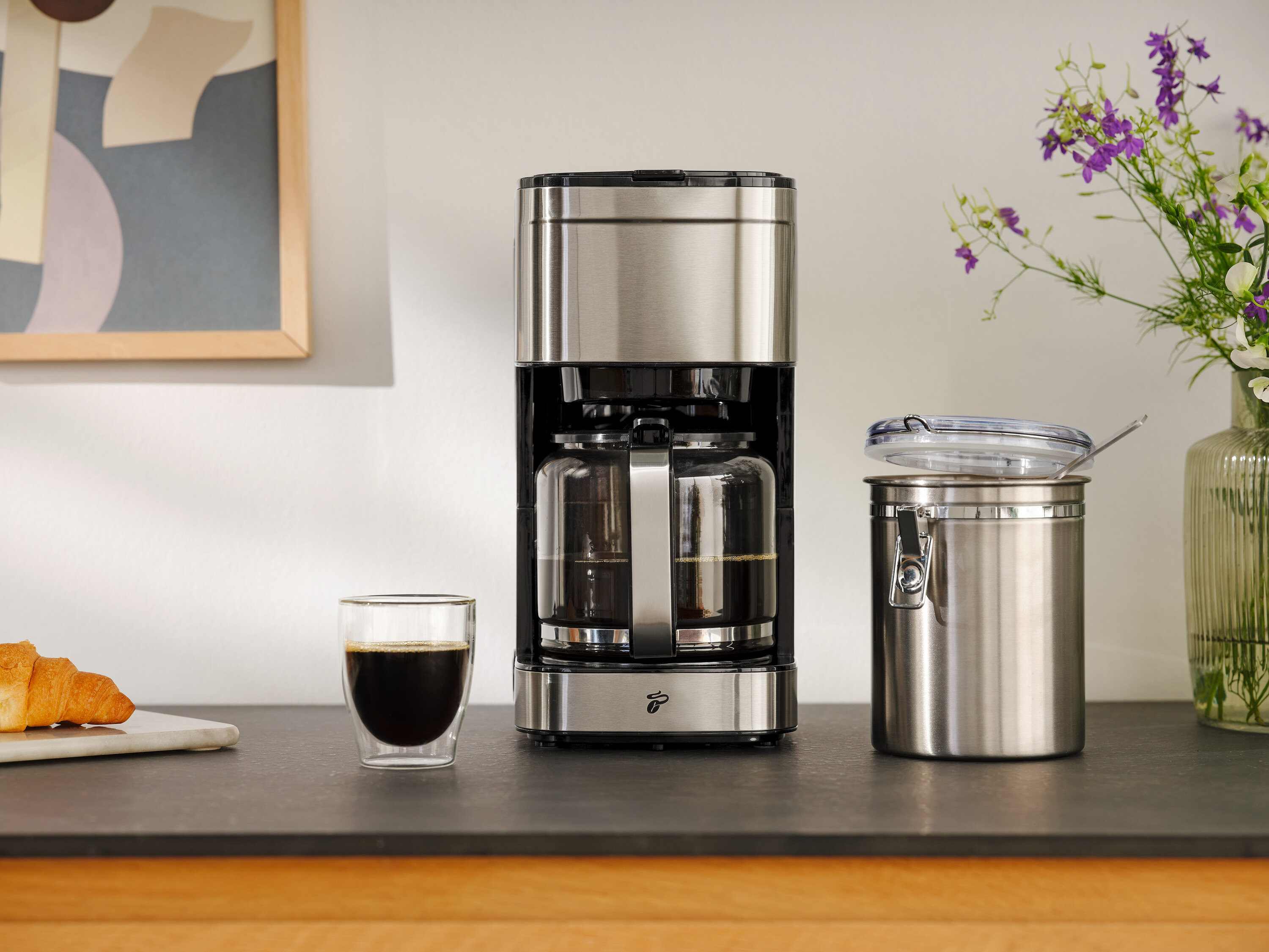 Kávovar vedle sklenice kávy, croissantu a skladovací nádoby.