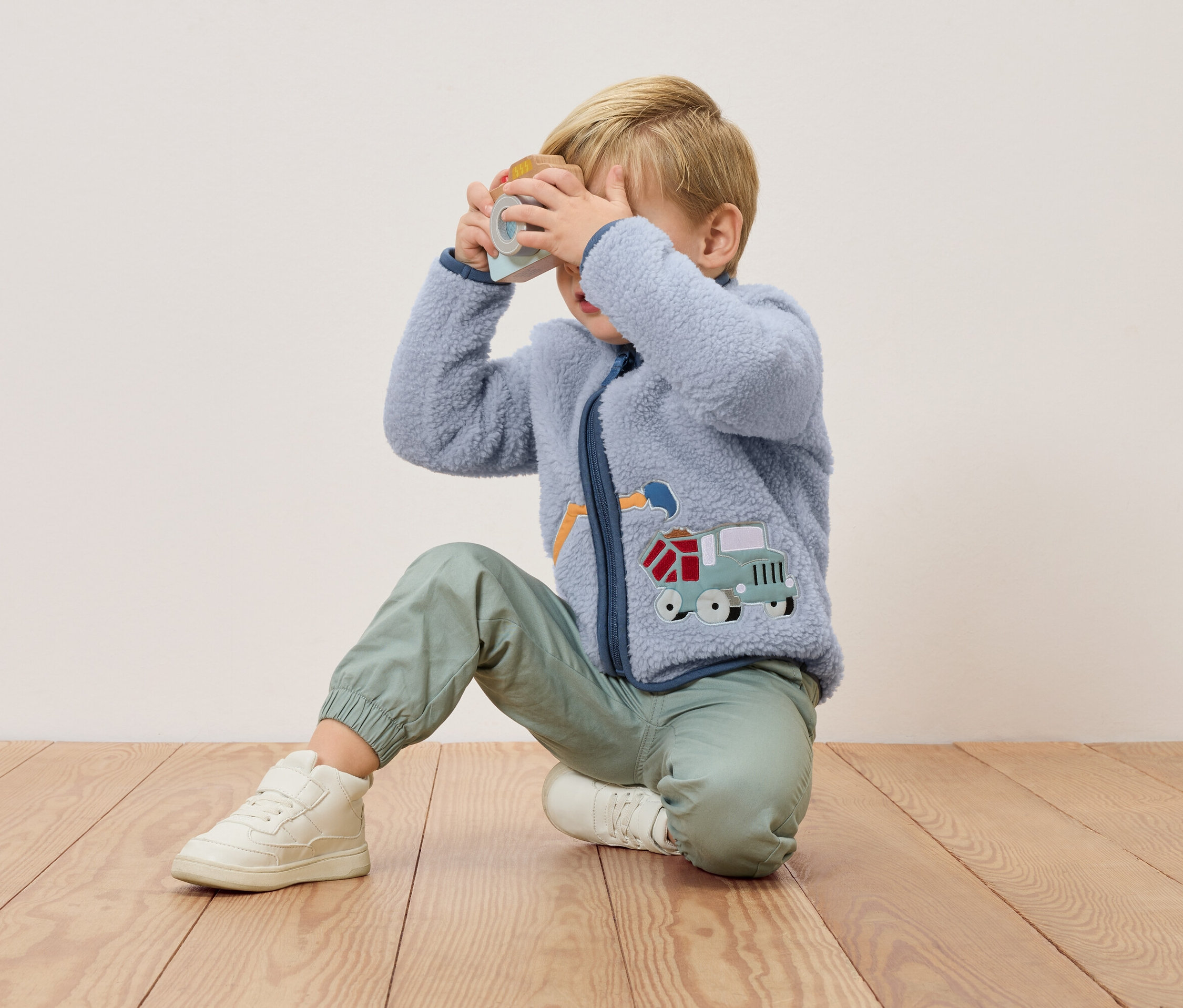 Blonďatý chlapec klečí na dřevěné podlaze a drží před obličejem hračkový fotoaparát. Má na sobě modrou fleecovou bundu s aplikací náklaďáku a zelené kalhoty.