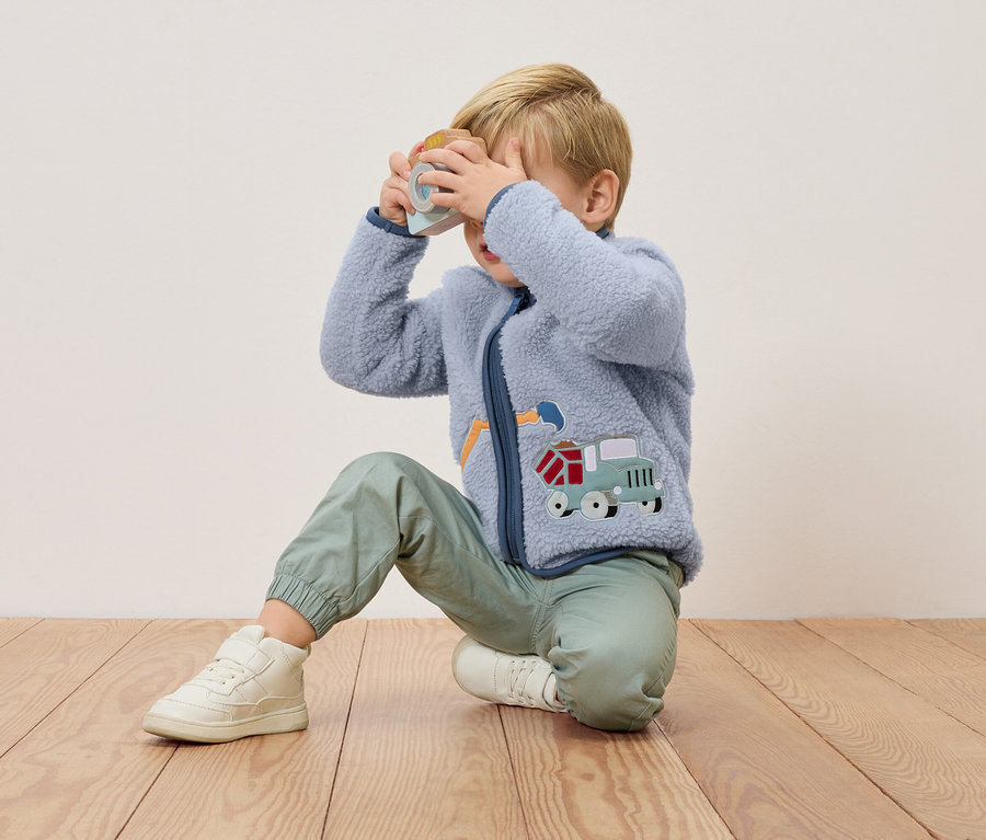 Blonďatý chlapec klečí na dřevěné podlaze a drží před obličejem hračkový fotoaparát. Má na sobě modrou fleecovou bundu s aplikací náklaďáku a zelené kalhoty.