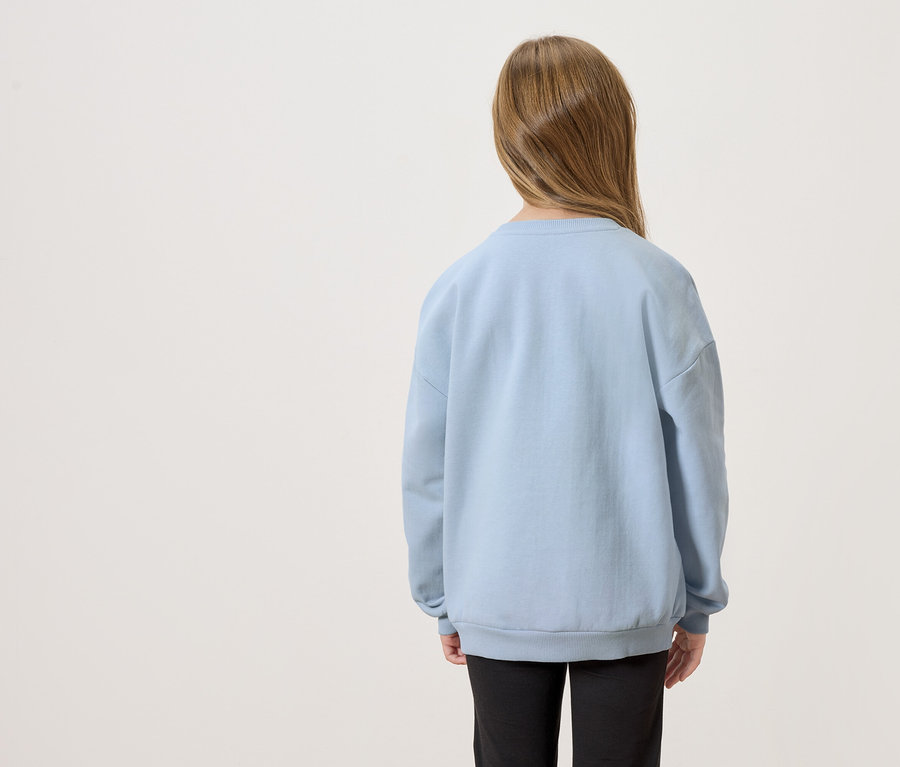 Pohled zezadu na dítě s dlouhými hnědými vlasy, oblečené ve světle modrém svetru a černých kalhotách.