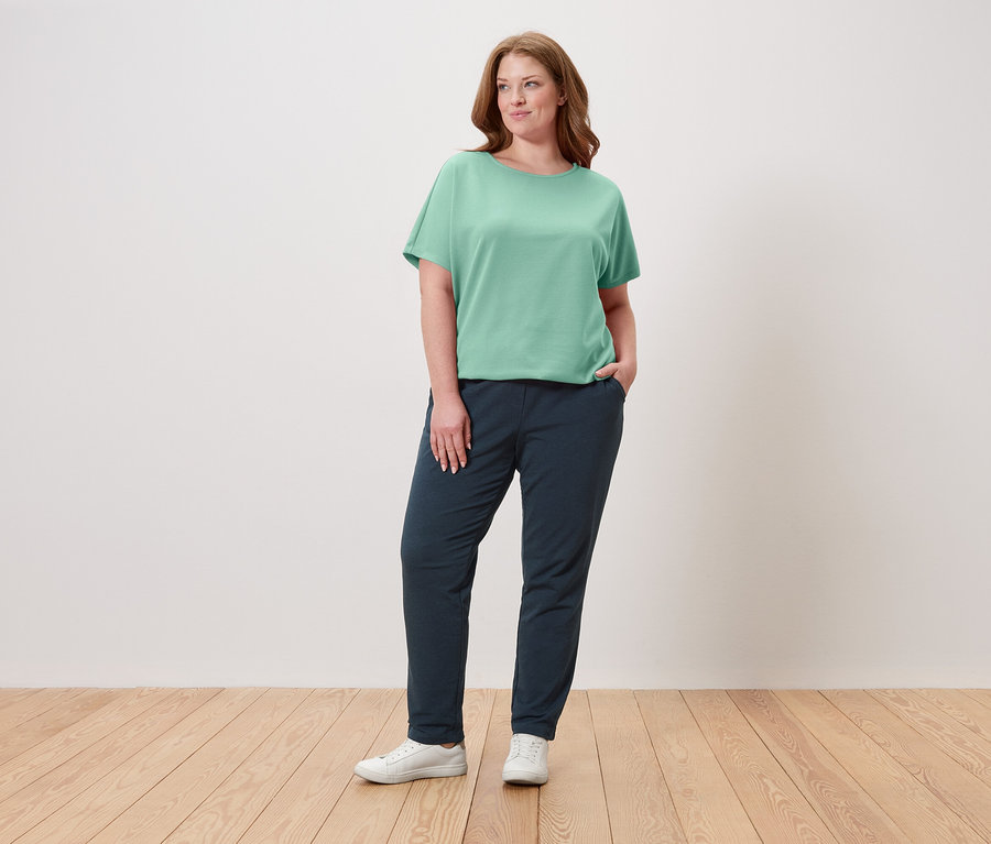 Žena stojí s rukou v kapse, má na sobě zelené tričko, tmavě modré kalhoty a bílé tenisky.
