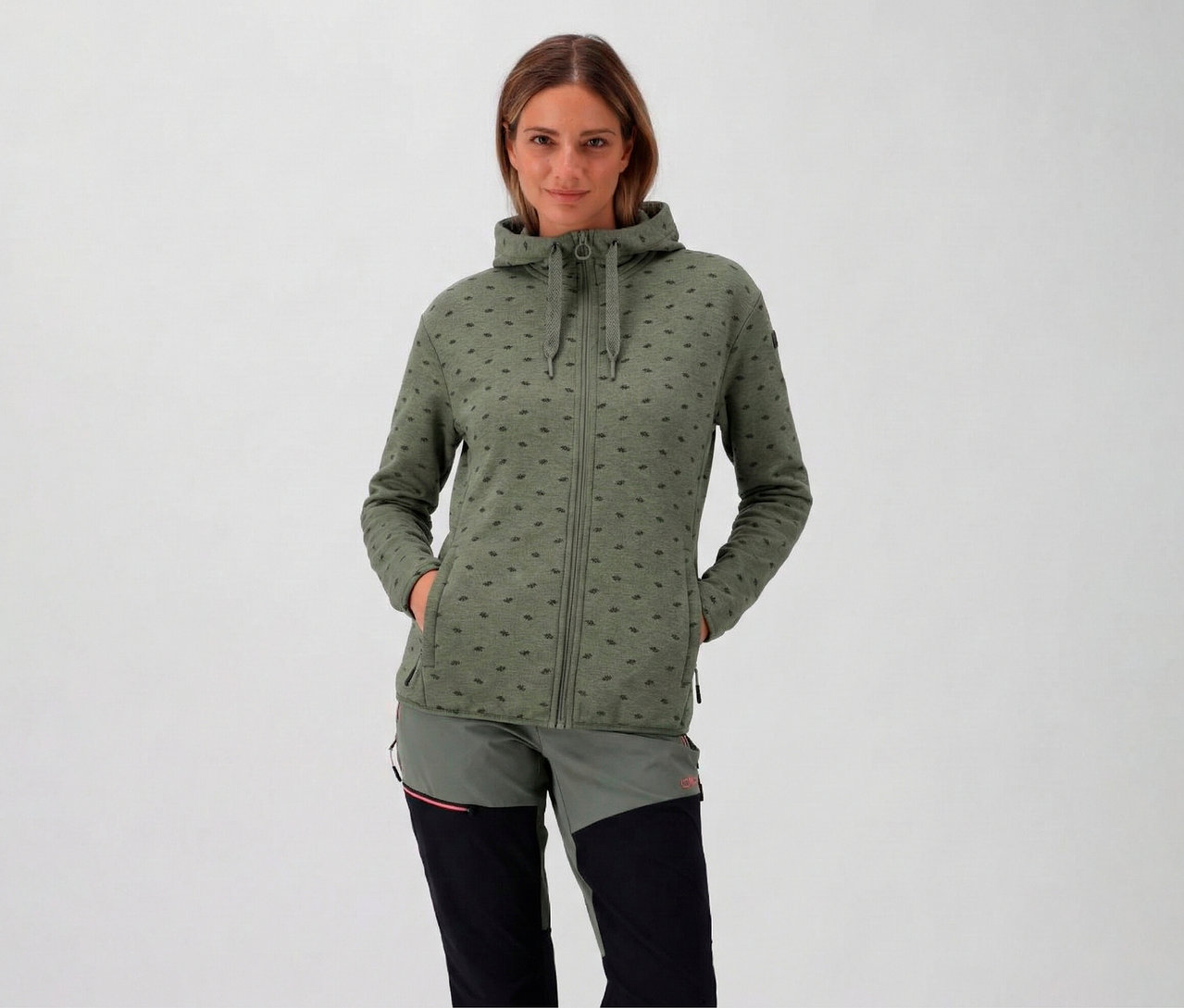 Žena s kapucí má na sobě dámskou fleecovou bundu s kapucí CMP, avokádová a má ruce v kapsách.