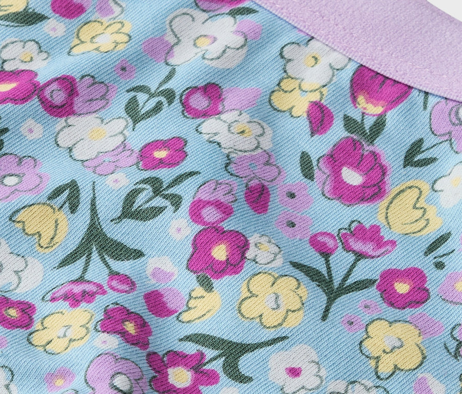 Detail látky s květinovým vzorem v růžové, žluté a bílé barvě na světle modrém pozadí a fialovou stuhou.