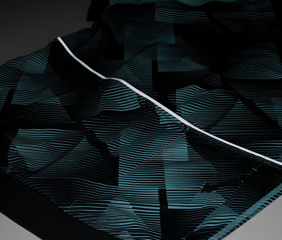 Detail oděvu s opakujícím se geometrickým vzorem v černé a tyrkysové barvě, zvýrazněný bílým pruhem.