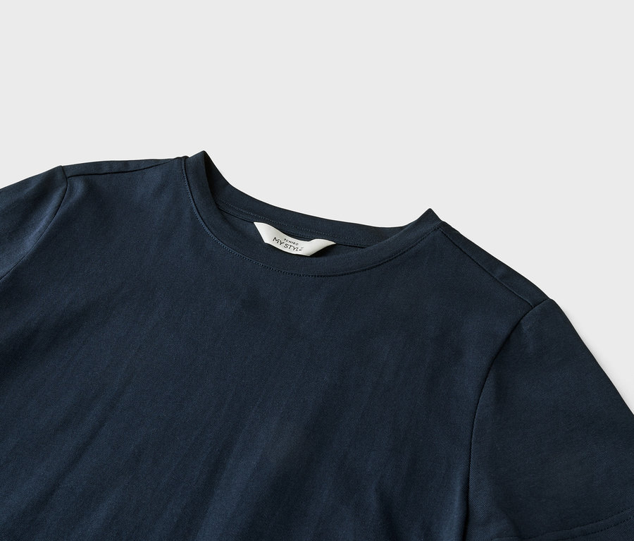 Detailní záběr na tmavě modré tričko s bílým štítkem s nápisem "ICHIBO MY STYLE" na límci, ležící na světle šedém pozadí.