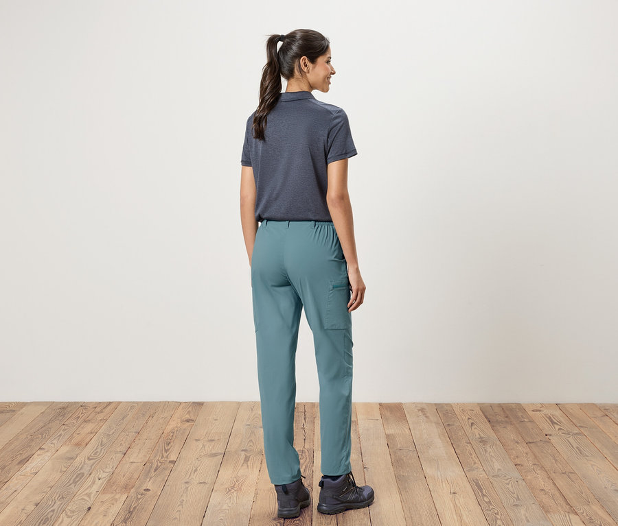 Pohled zezadu na ženu s culíkem, oblečenou v tmavě modré polokošili, zelených kalhotách a trekových botách, stojící před bílou stěnou a dřevěnou podlahou.