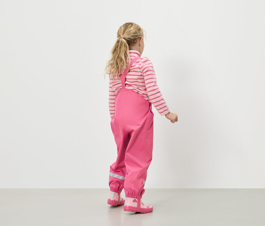 Pohled zezadu na dívku s blond vlasy, oblečenou do růžovo-bílého pruhovaného svetru, růžových dešťových kalhot s laclem a růžových gumáků se srdíčky.