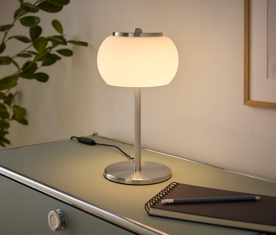 Svítící stolní lampa s bílým kulovým stínítkem a kartáčovanou kovovou základnou stojí na světle zelené komodě. Vedle ní leží zápisník a tužka. Vlevo je rostlina, vpravo zarámovaný obraz na zdi.