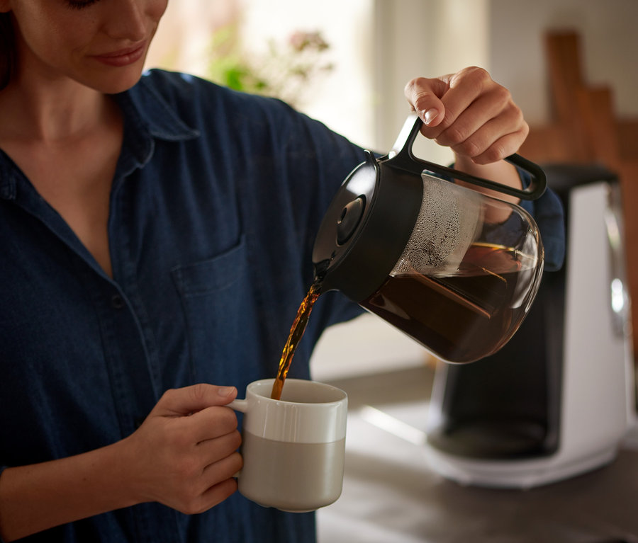 Žena nalévá kávu z konvice do hrnku. V pozadí je vidět překapávač na kávu »Let's Brew«.