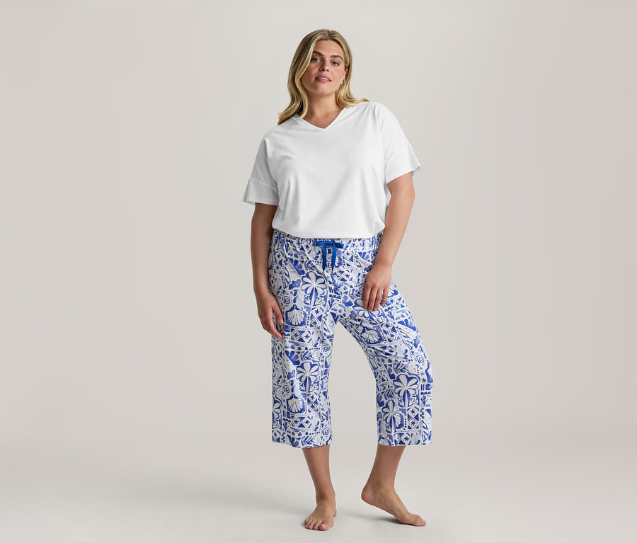 Žena stojí, oblečená v bílém tričku s V-výstřihem a modro-bílých vzorovaných 3/4-Pyjama-Set kalhotách s modrou šňůrkou, bosá.