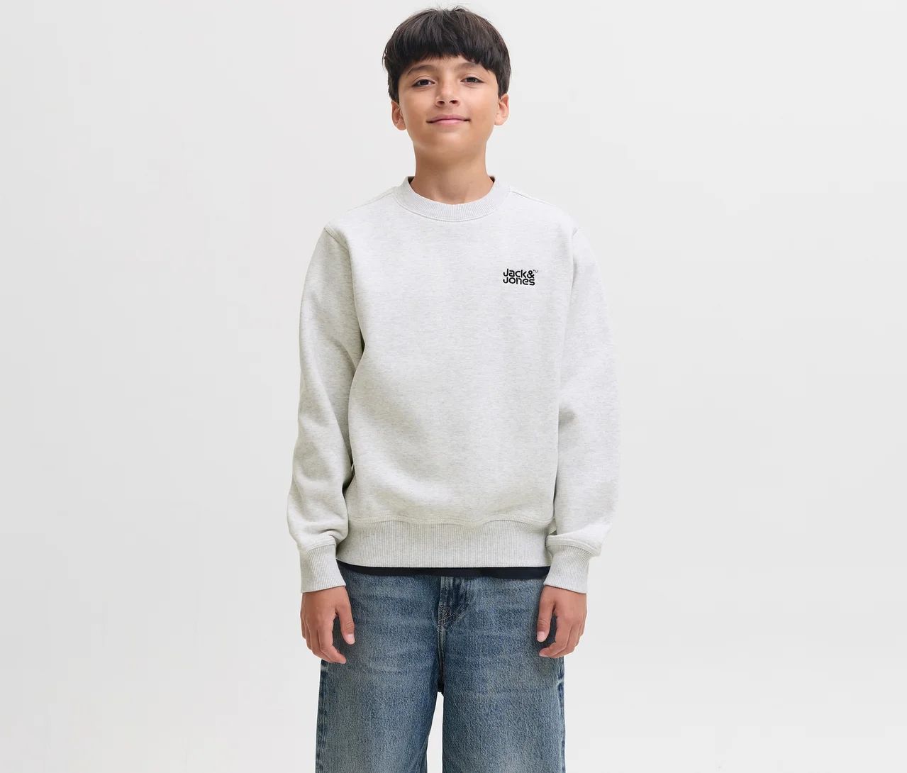 Mladý chlapec s tmavými vlasy se mírně usmívá, má na sobě bílou mikinu Jack & Jones Colton s logem značky na hrudi a modré džíny.