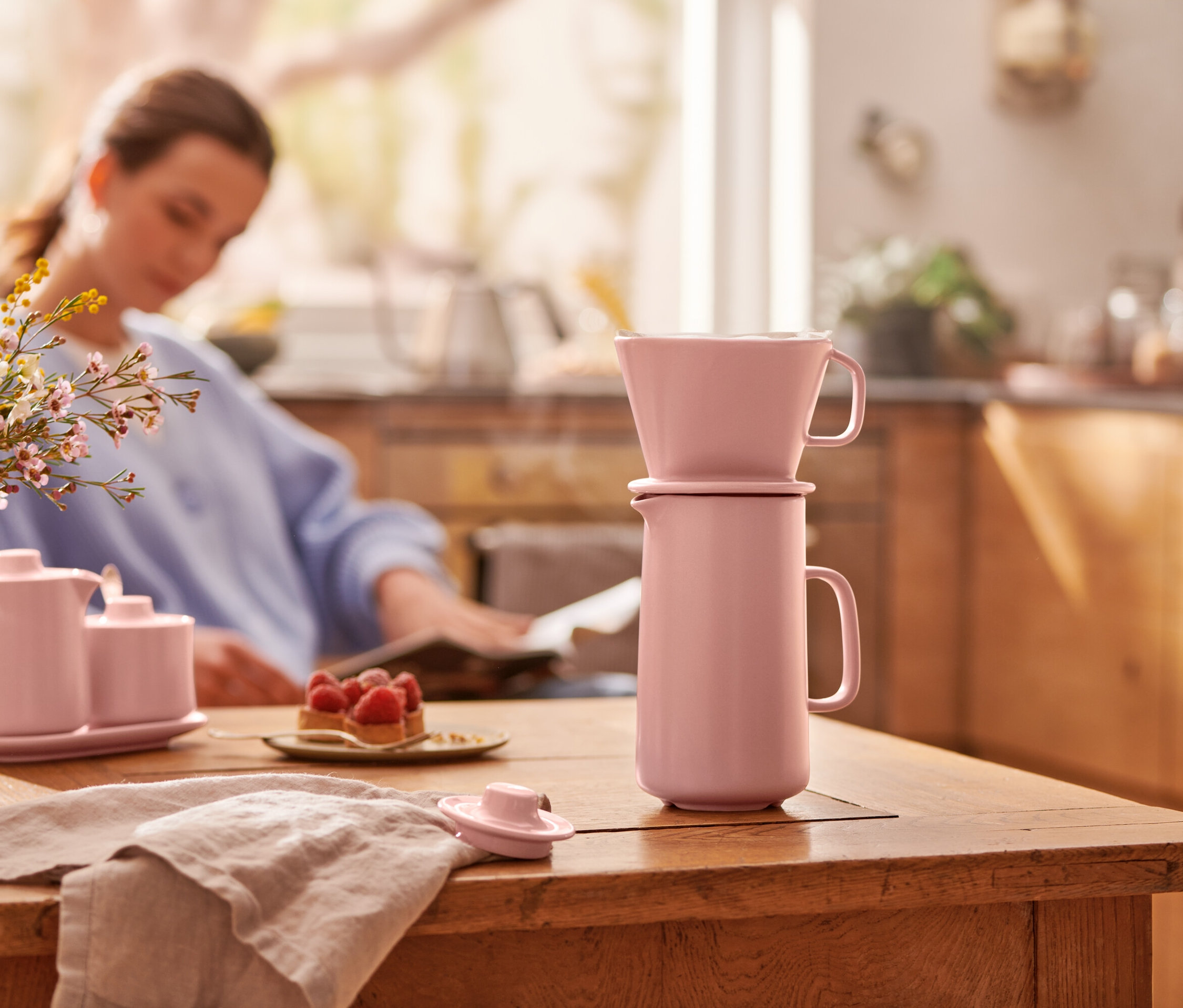 Růžová konvička na kávu s filtrem stojí na dřevěném stole. V pozadí sedí žena.