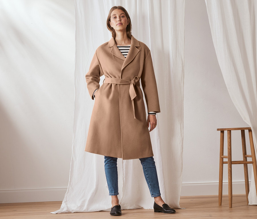 Žena pózuje ve vlněném kabátě s kašmírem světle hnědé barvy, s páskem. Kabát má na sobě přes proužkované tričko a džíny.