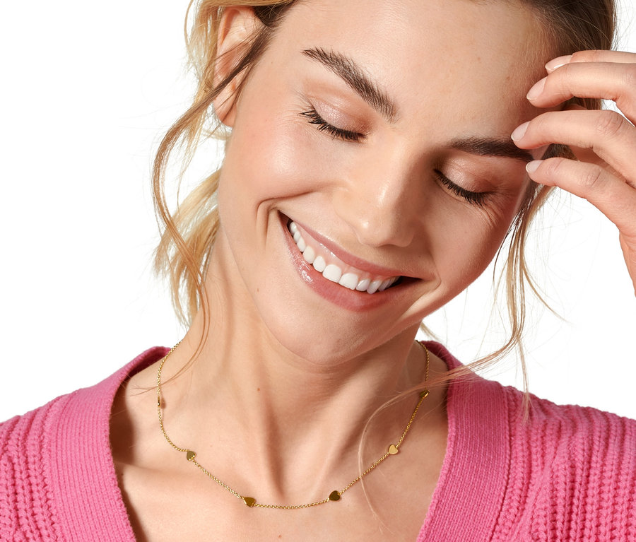 Usmívající se žena se zavřenýma očima se dotýká obličeje a má na sobě vrstvený náhrdelník »Happy«.